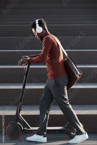 Side view of young African man listening to music in headphones while pushing scooter against staircase photo