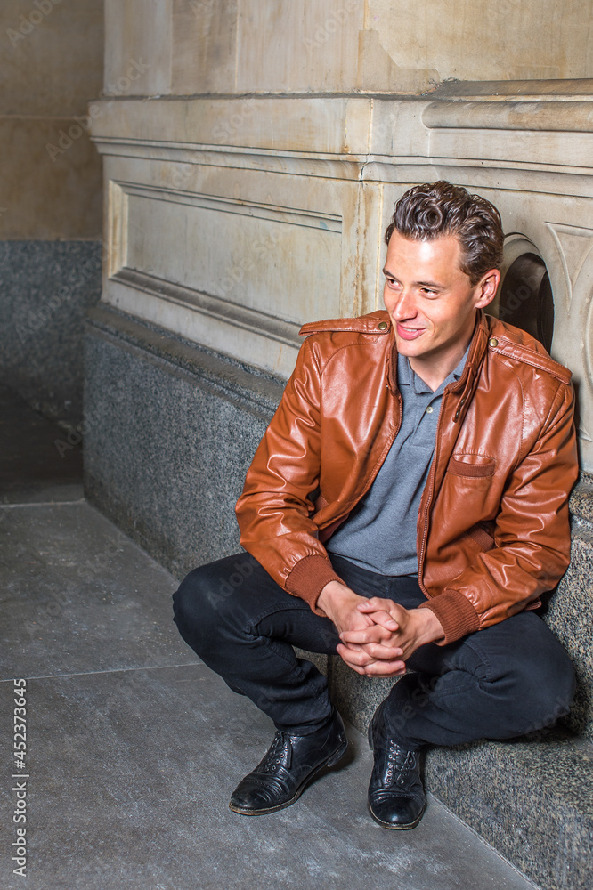 Dressing in a brown leather jacket, gray undershirt, black pants, holding hands, a young handsome guy is squatting in the corner of a building, relaxing and thinking.. Dressing in a brown leather jacket, gray undershirt, black pants, holding hands, a young handsome guy is squatting in the corner of a building, relaxing and thinking..