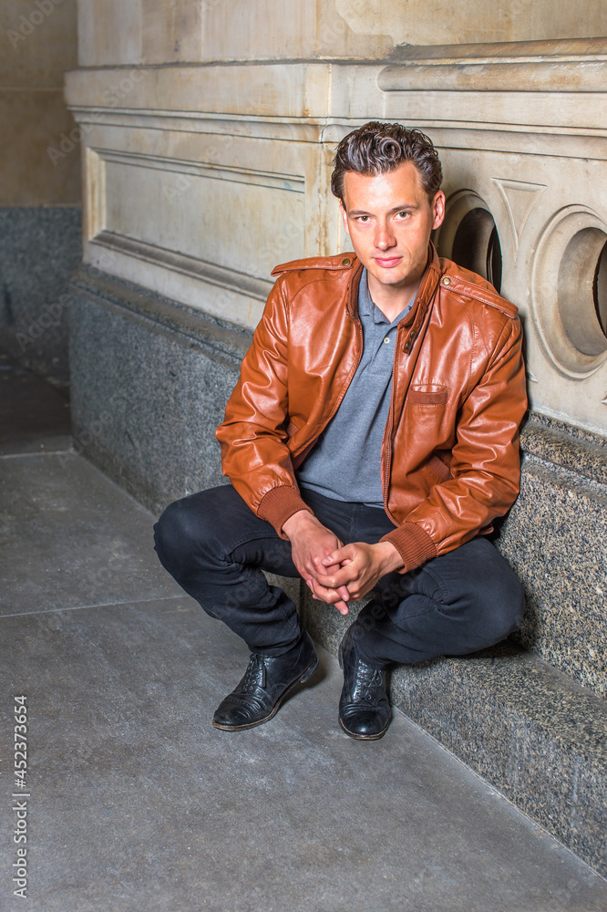Dressing in a brown leather jacket, gray undershirt, black pants, holding hands, a young handsome guy is squatting in the corner of a building, relaxing and thinking... Dressing in a brown leather jacket, gray undershirt, black pants, holding hands, a young handsome guy is squatting in the corner of a building, relaxing and thinking...