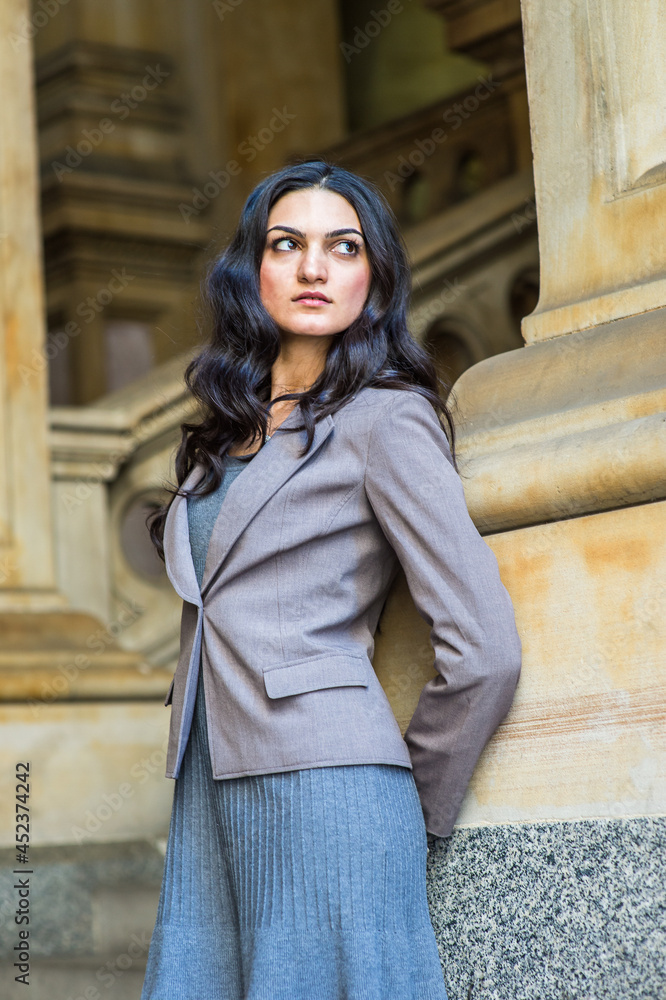 Dressing in a gray knit under dress and a jacket, a young businesswoman is standing outside an office building, hands putting in back, leaning against the wall, confidently looking forward. Dressing in a gray knit under dress and a jacket, a young businesswoman is standing outside an office building, hands putting in back, leaning against the wall, confidently looking forward.
