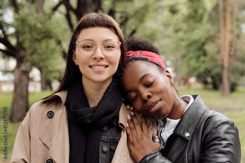 Happy young woman in eyeglasses looking at camera while her girlfriend keeping head on her shoulder