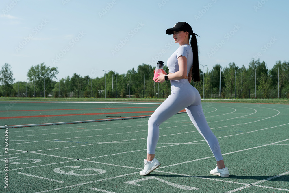 An Asian track and field athlete in a sports gray suit stands at the stadium with her left foot forward and holds a pink water bottle in her hand