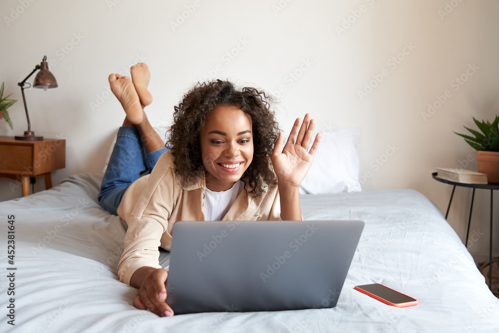 Young African American woman lying on bed having virtual meeting waving hand chatting with friends during online video call at home having fun of friendly casual communication using laptop computer. Young African American woman lying on bed having virtual meeting waving hand chatting with friends during online video call at home having fun of friendly casual communication using laptop computer.