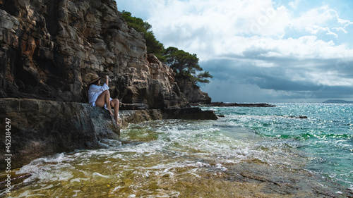 Attractive brunette sitting on a rock on the shores of the Adriatic sea , island of Vis in Croatia. Cloudy summer day, beautiful teal sea in the distance, white waves hitting the rocks