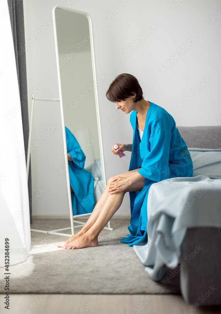 Obraz premium Attractive mature woman in blue dressing gown sitting on bed and applying moisturising cream on legs. Concept of skin care and beauty routine.