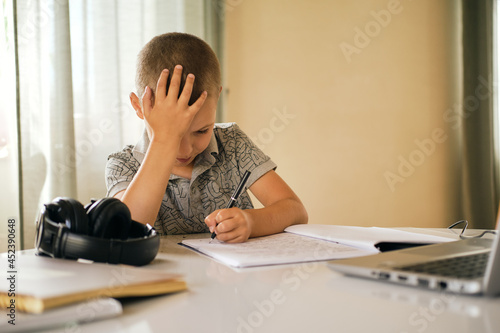 School boy writing in notebook and using laptop while doing schoolwork at home or during online lesson. Online education concept.