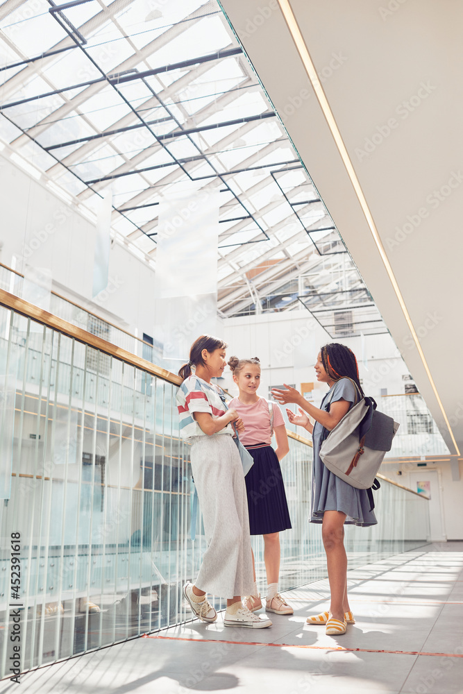 Vertical full length portrait of three schoolgirls chatting in school while standing in sunlight under glass ceiling, copy space