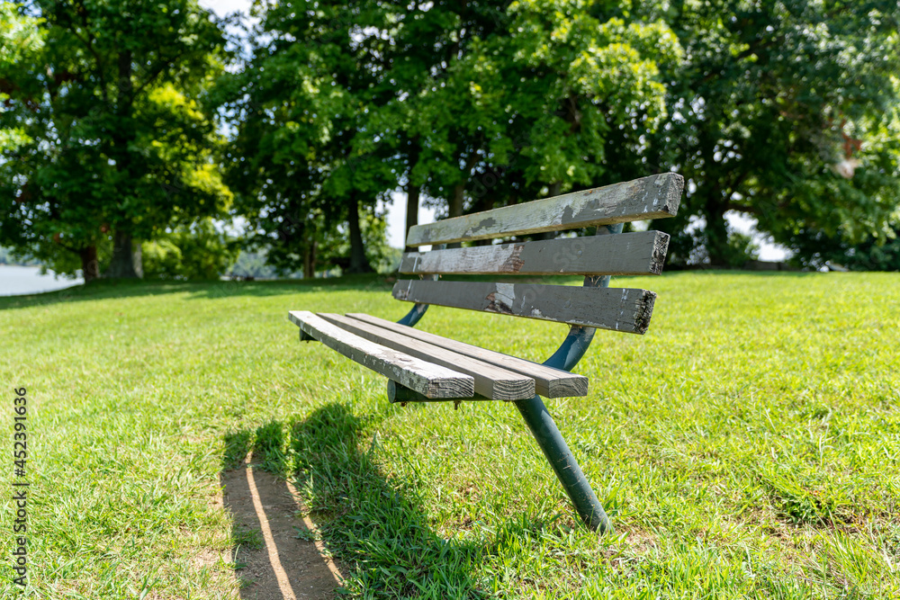 An old deteriorating wood and metal park bench in a grass field on a sunny summer day surrounded by tall trees. An old deteriorating wood and metal park bench in a grass field on a sunny summer day surrounded by tall trees.