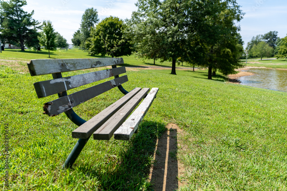 An old deteriorating wood and metal park bench in a grass field on a sunny summer day surrounded by tall trees. An old deteriorating wood and metal park bench in a grass field on a sunny summer day surrounded by tall trees.