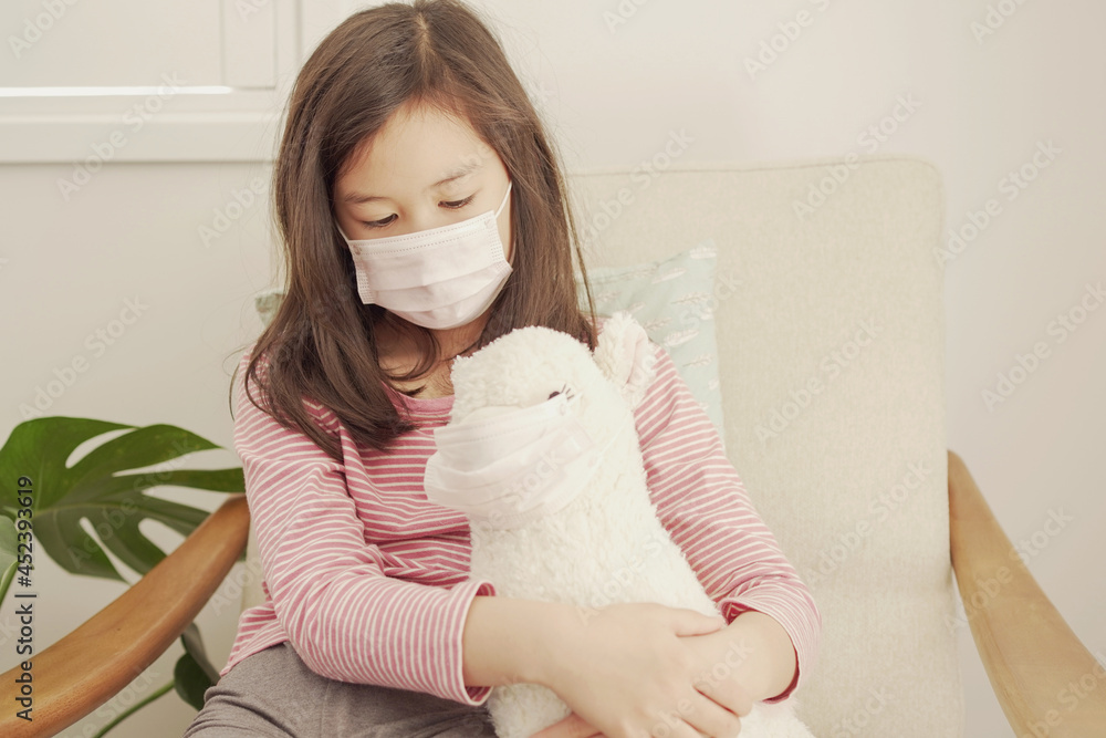 Mixed Asian girl wearing face mark hugging her plush toy at home, social distancing, unwell kid staying at home, child health concept Mixed Asian girl wearing face mark hugging her plush toy at home, social distancing, unwell kid staying at home, child health concept