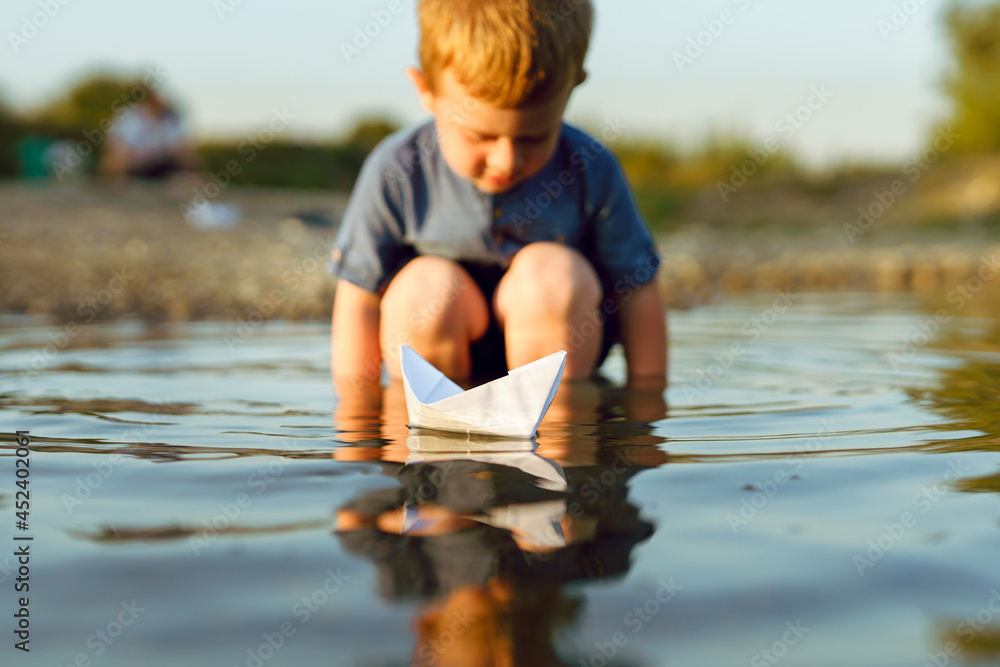 Fototapeta premium Small caucasian boy blonde hair three years old sitting alone on the coast of the river or lake in sunny summer or autumn evening playing with paper boat childhood tranquility concept copy space