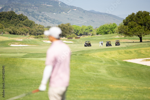 A view of golf carts parked in the distance grassy field, while a golfer stand in the foreground in bokeh, seen at a local golf course.