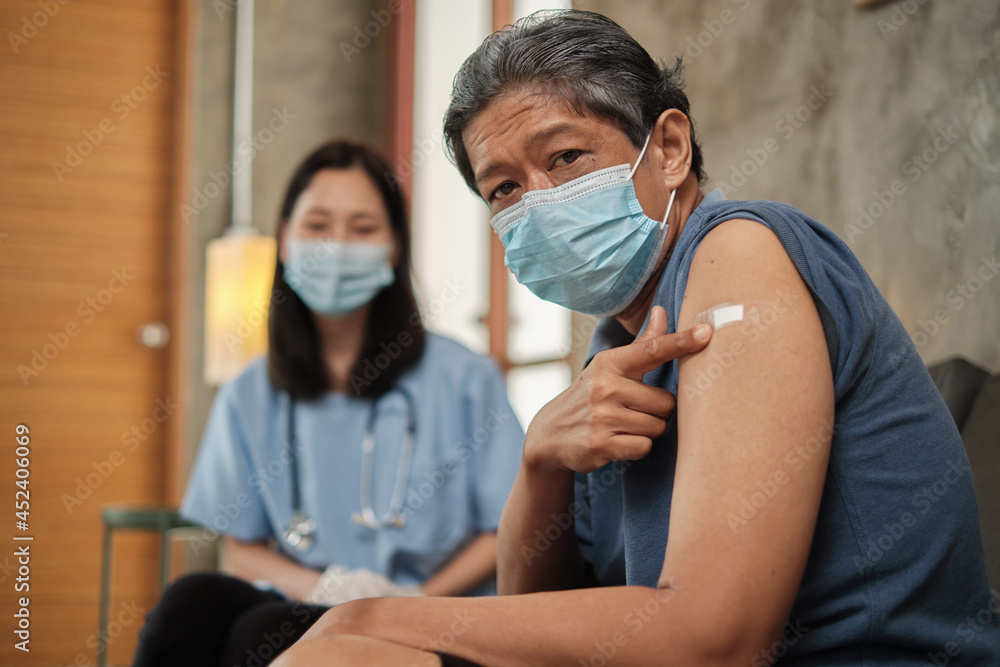 Elderly Asian male patient points a finger at a plaster-covered on his arm after vaccinating against COVID-19. This is a medical service from a specialist doctor at a retirement home.