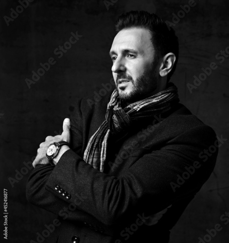 Black and white. Elegant bearded man in jacket and stylish scarf stands sideways, holding arms crossed at chest and looks right ahead over dark background