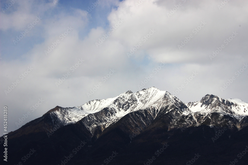 a dark mountain range with snow-capped peaks against a background of white clouds