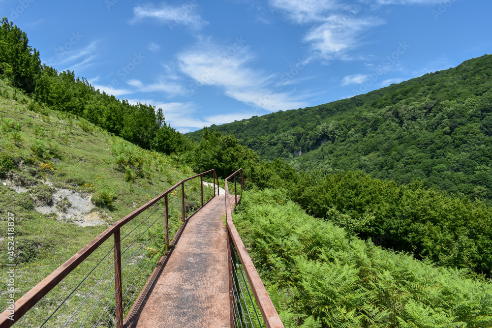 Hiking trail leading to Okatse Canyon in village Gordi, Imereti region, Georgia. Path surrounded by hills covered by green trees. Blue sky with light clouds above. Georgian tourist attraction