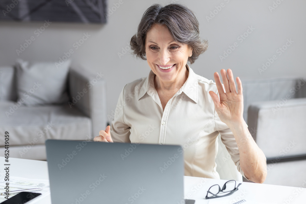 Caucasian aged business woman waves hand, greets the participants in the video conference, used laptop computer to communicate online sitting in the office or at home. Video call, remote work concept Caucasian aged business woman waves hand, greets the participants in the video conference, used laptop computer to communicate online sitting in the office or at home. Video call, remote work concept