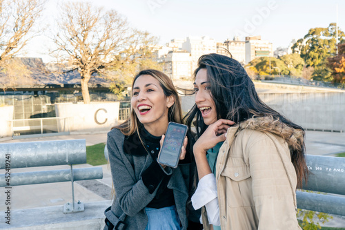 Two beautiful Latin women sharing cell phone messages while laughing in a public park. Technology and communications concept.