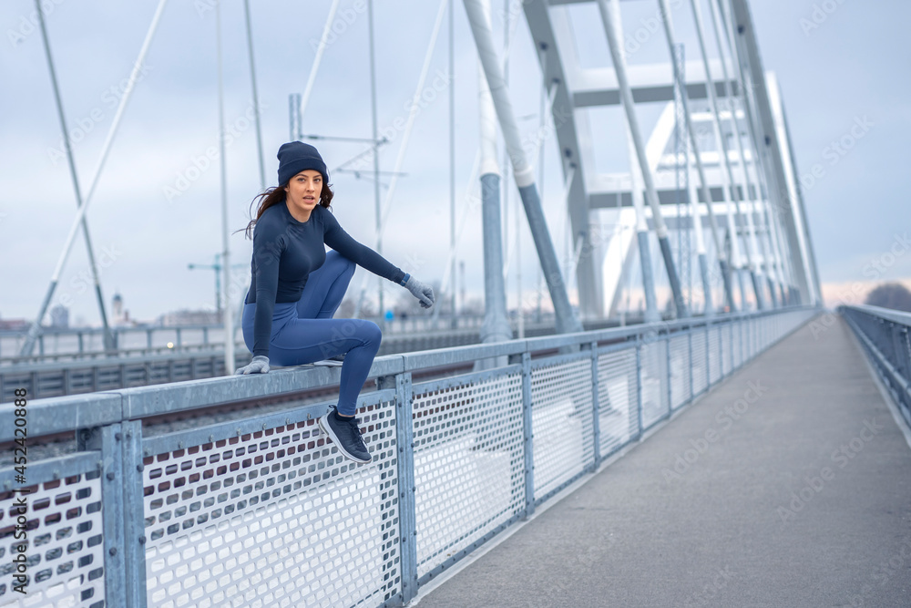 Young happy fitness woman in winter blue sportswear skips over bridge fence during cold winter day