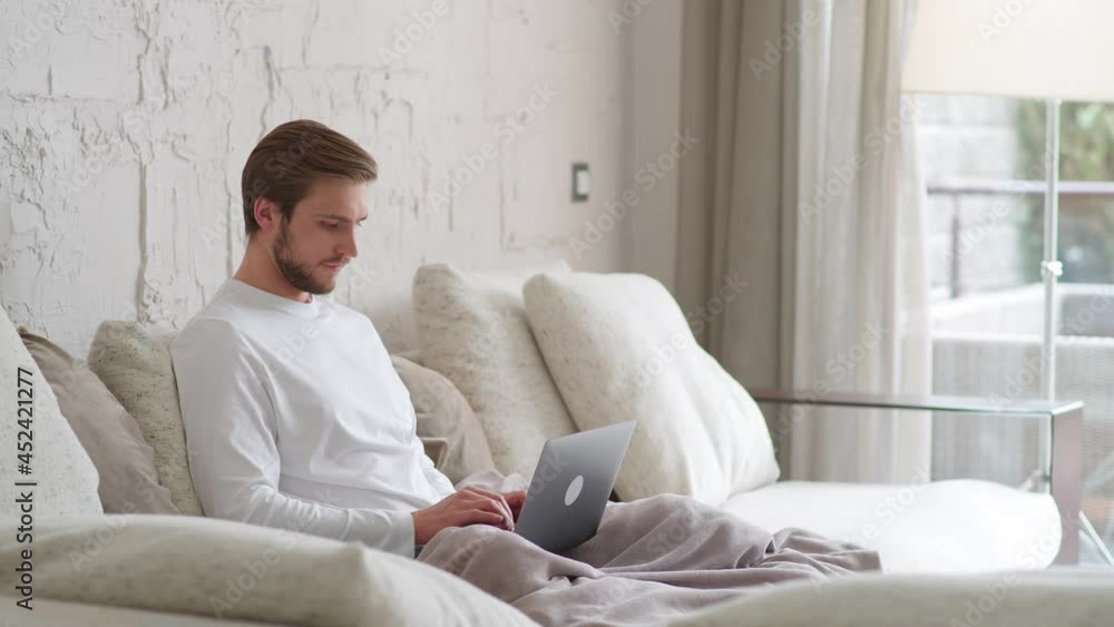 Cheerful caucasian man sits at home on the couch and works using a laptop, financial manager on a remote work during self-isolation in quarantine, a man makes good deal, positive emotions, 4k slow