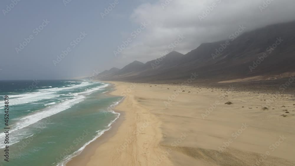 aerial drone shot of big waves crashing on the long white beach of cofete with beautiful mountains in the background in Fuerteventura, canary island. aerial drone shot of big waves crashing on the long white beach of cofete with beautiful mountains in the background in Fuerteventura, canary island.