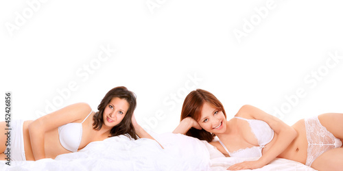 Two slim young women wearing white underwear, they are smiling and lying on a bed in front of white background