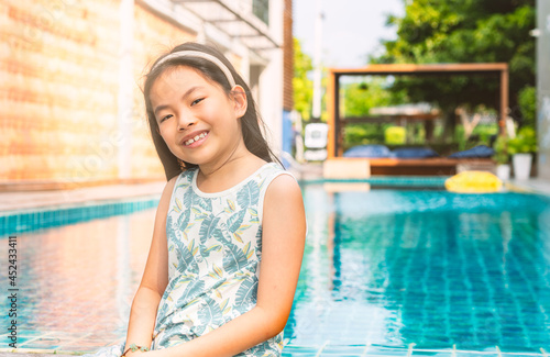 Portrait Asian little girl at swimming pool, casual cute dress, smiling face, blurred background of swimming pool at a resort. © dul_ny