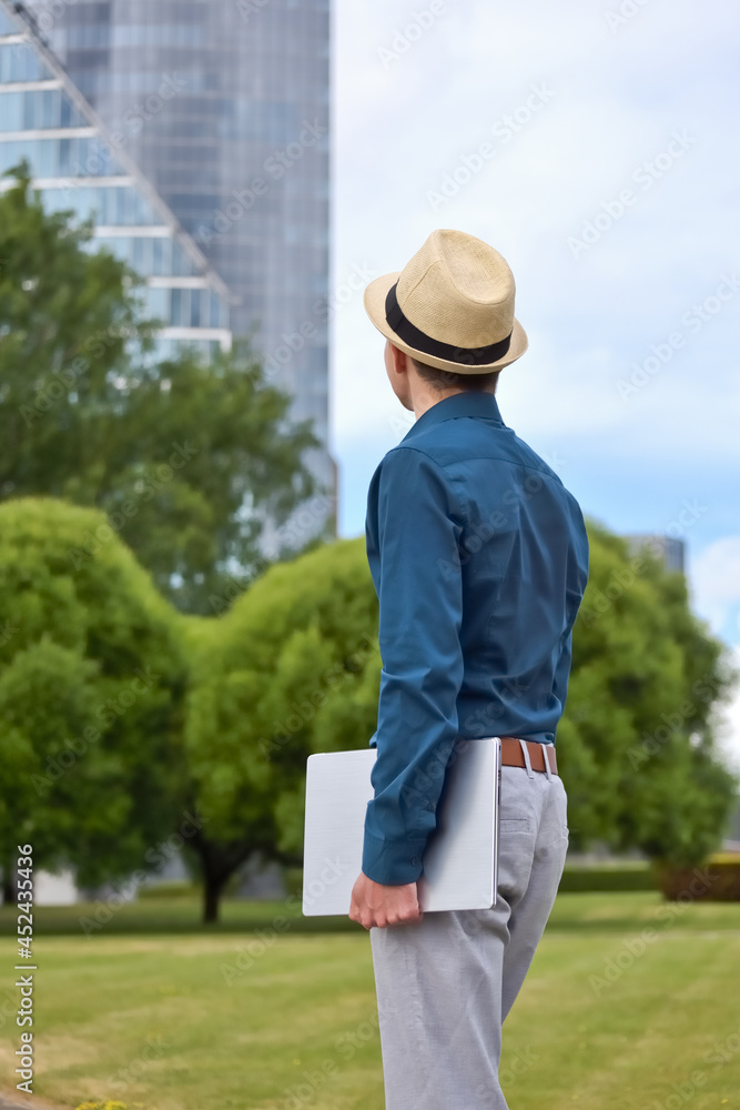 A man with a laptop in his hand stands and looks towards the office building. Young student, programmer and businessman with computer in hand looks at office building and thinks about work and career.
