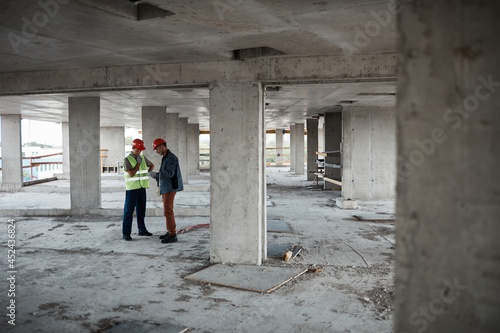 Wide angle view at two building contractors discussing project while standing at construction site, copy space