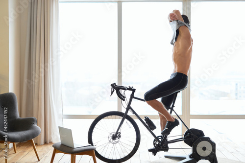 Athletic young man taking off shirt while cycling indoors at home