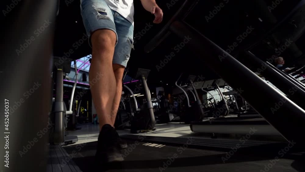 the legs of a sporty man in torn denim shorts. he's running on a treadmill. low angle of view the legs of a sporty man in torn denim shorts. he's running on a treadmill. low angle of view