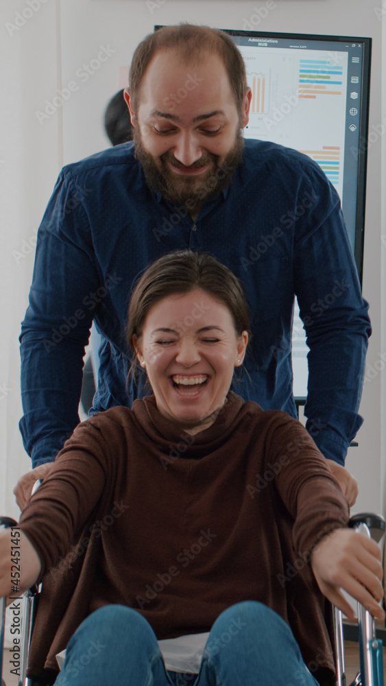 Cheerful employee taking break pushing wheelchair with his paralysed invalid disabled colleague smiling and having fun in start up financial business office. Immobilized woman laughing in workplace Cheerful employee taking break pushing wheelchair with his paralysed invalid disabled colleague smiling and having fun in start up financial business office. Immobilized woman laughing in workplace