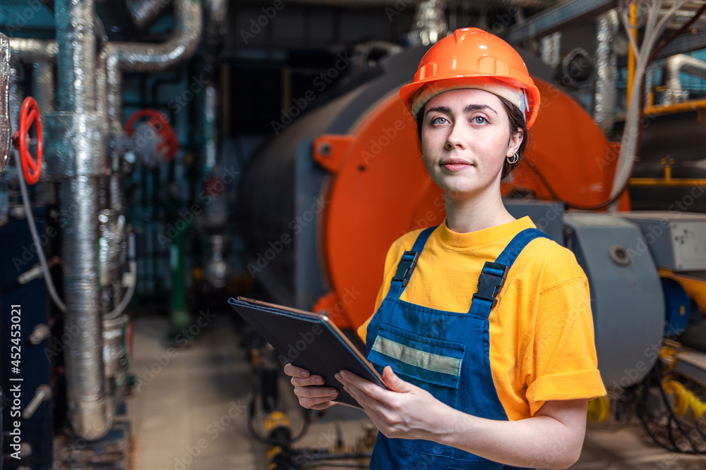 Portrait of a young worker in a helmet and uniform, with a tablet in her hand. In the background is a boiler room. Concept of industrial production