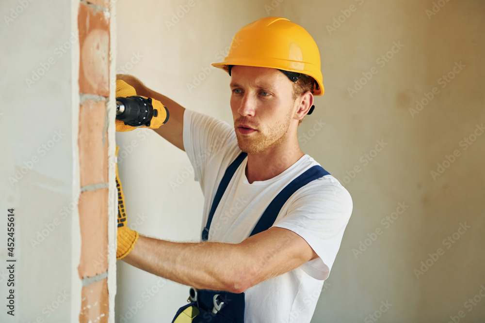 Indoors in unfinished room. Young man working in uniform at construction at daytime Indoors in unfinished room. Young man working in uniform at construction at daytime