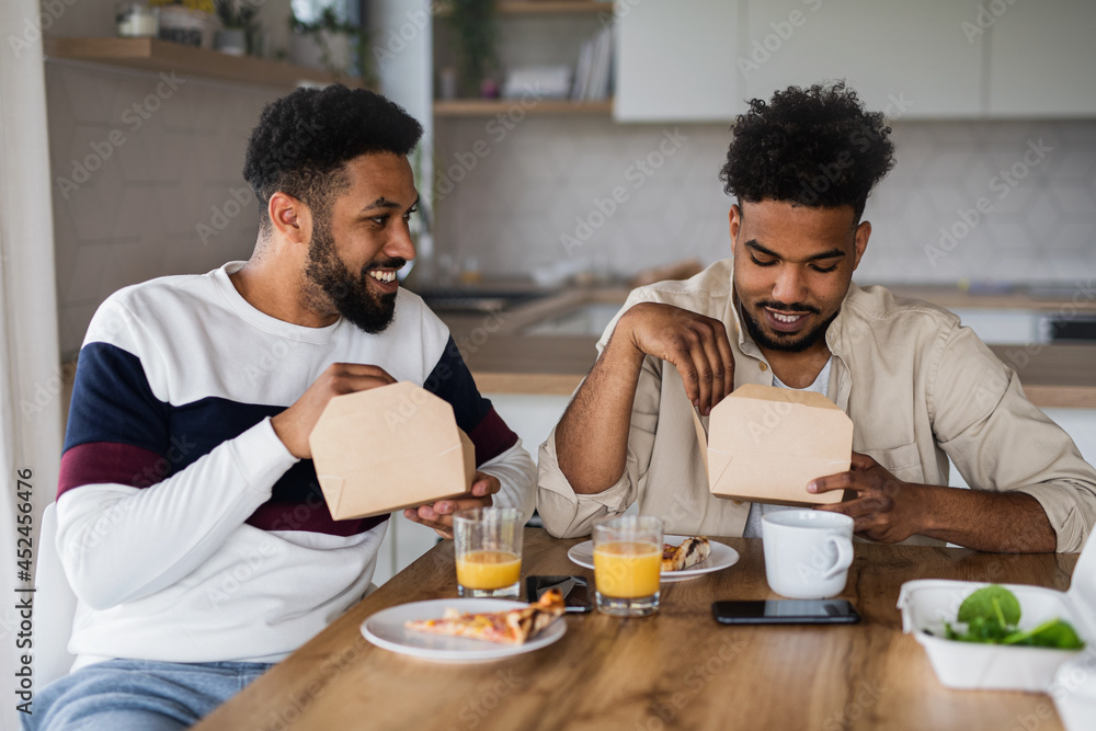 Portrait of young adult brothers sitting in kitchen indoors at home, eating take away food. Portrait of young adult brothers sitting in kitchen indoors at home, eating take away food.