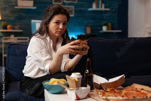 Portrait of relaxed woman looking into camera holding tasty burger in hands during fast food night in living room. Caucasian female enjoying delicious home delivered junk-food. Takeaway delivery