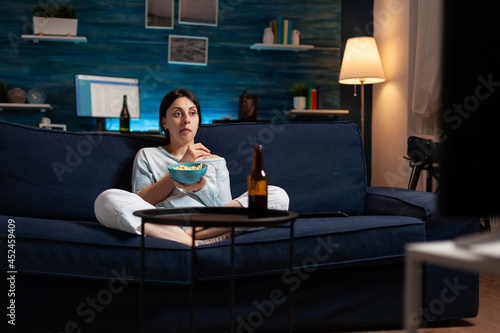 Interested woman eating popcorn and watching entertainment serial on tv. Focused home alone at night with surprised face looking at suspense movie sitting on comfortable interested couch. photo