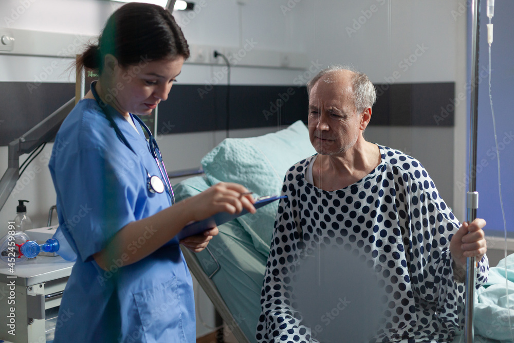 Obraz premium Senior sick man in hospital room discussing with nurse about treatment during consultation. Medical staff with stethoscope writing notes on clipboard preparing patient for surgery.