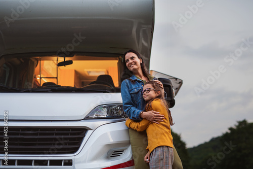 Mother with daughter standing by car outdoors in campsite at dusk, caravan family holiday trip.