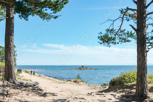 Ground back road along coast of calm blue sea with little island at summer sunny day, beautiful scenic landscape of walking vacation on lake Baikal, shore stroll away, natural frame by pine trees © Clara_Sh.