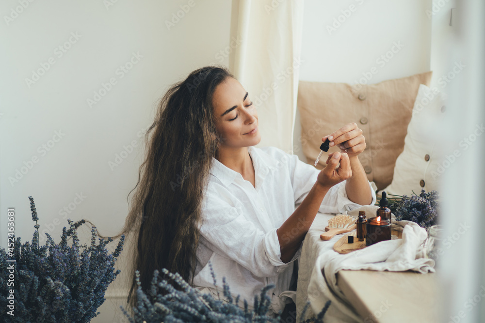 Young woman applying natural organic essential oil on hair and skin. Home spa and beauty rituals. - obrazy, fototapety, plakaty 