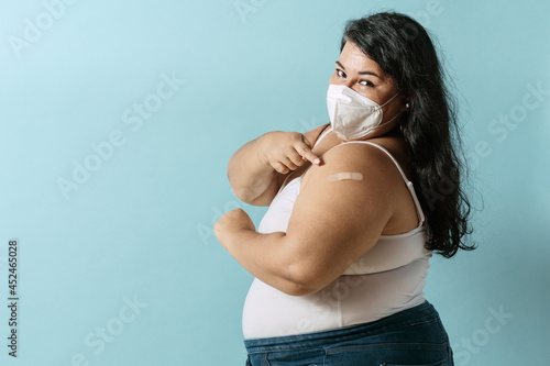 Oversized latinx woman using face mask pointing with her finger an bandage vacci Fotobehang