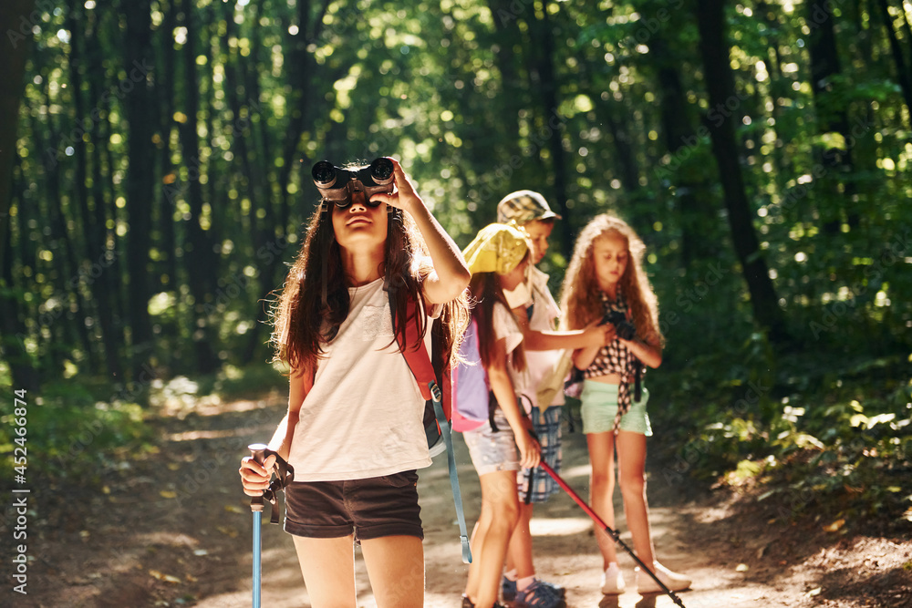 Girl standing in front of her friends. Kids strolling in the forest with travel equipment Girl standing in front of her friends. Kids strolling in the forest with travel equipment