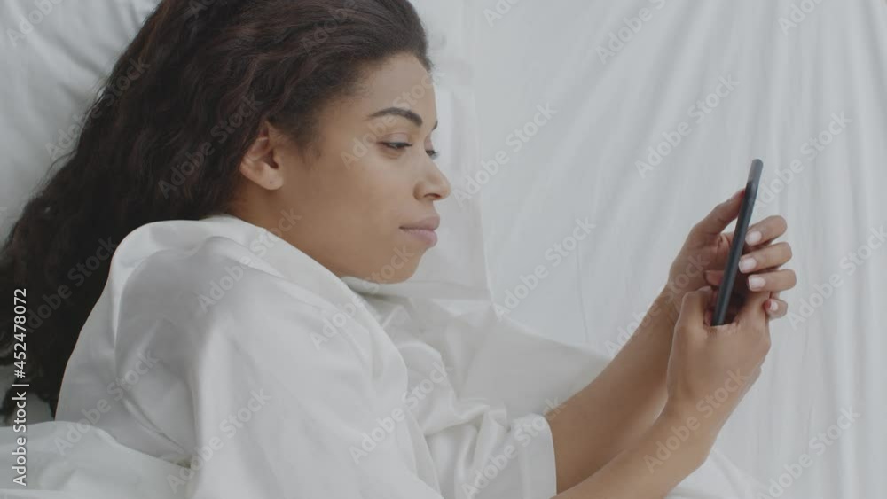 Young african american woman web surfing social media on smartphone, lying in bed, profile portrait, top view