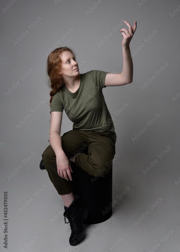 Full length portrait of pretty red haired woman wearing army green khaki shirt, utilitarian pants and boots. Sitting pose isolated on studio background.