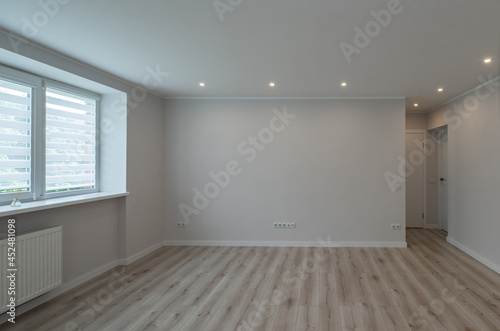 Modern light interior of empty renovated room with white walls. Heating battery under window. Beige parquet floor.