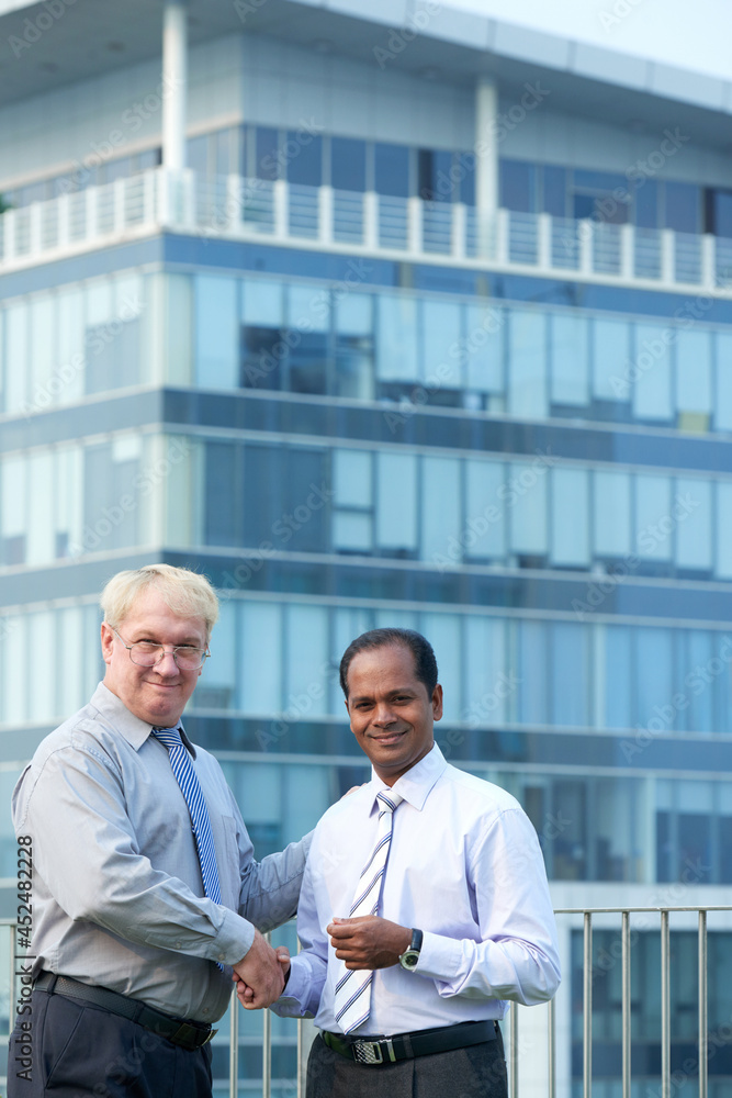 Diverse business partners standing on balcony of office building and shaking hands to confirm deal Diverse business partners standing on balcony of office building and shaking hands to confirm deal