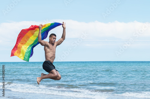 Young man holding a gay pride flag while jumping at the beach.