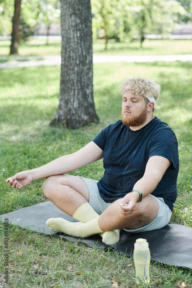 Overweight man sitting on the grass with eyes closed and concentrating on yoga exercises in the park Overweight man sitting on the grass with eyes closed and concentrating on yoga exercises in the park