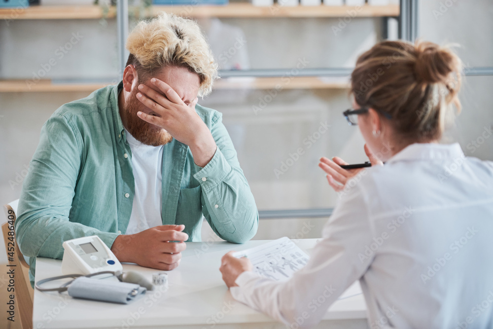 Upset overweight man sitting at the table and covering his face with hand he listening to diagnosis of the doctor at hospital